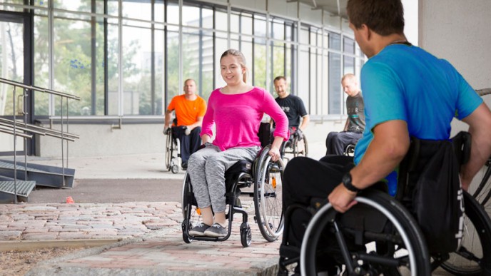 Nuori nainen harjoittelee pyörätuolilla liikkumista ja apuvälinekummi neuvoo vierestä.