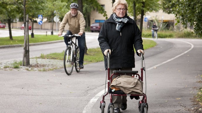 Vanhempi vaaleahiuksinen nainen mustassa takissa rollaattorin kanssa jalkakäytävän puolella yhdistetyllä pyörätiellä ja jalkakäytävällä. Pyörätien puolella ajaa takana vanhempi mies pyörällä pyöräilykypärä päässä. 