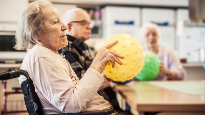 Etualalla vanha nainen pyörätuolissa pöydän äärellä keltainen jumppapallo kädessä, taustalla myös vanha mies ja nainen. Naisella vihreä pallo kädessä