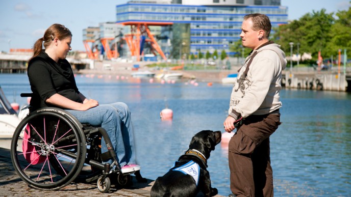 Nainen pyörätuolissa, avustajakoira ja mies ovat rannalla, vastarannalla rakennus.