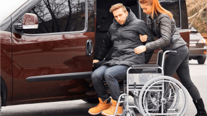 Naispuolinen henkilökohtainen avustaja auttaa pyörätuolissa istuvaa miestä autoon. Kuvituskuva.
