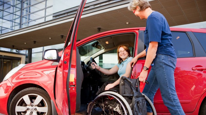 Punainen auto jossa nainen kuljettajana. Mies tuo pyörätuolin auton eteen. Kuvituskuva.