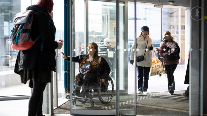 Ihmisiä kulkemassa lasiovista, henkilö pyörätuolilla pyöröovien keskellä